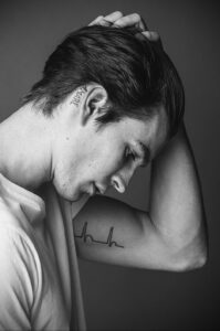 Черно-белое фото татуировок на шее и предплечье мужчины. Имя 'MARIE' за ухом и линия кардиограммы на руке.