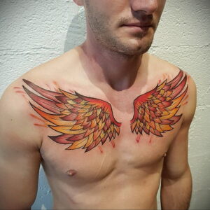 Фото тату огненных крыльев на мужской груди. Яркие оранжевые, желтые и красные цвета, детальная проработка перьев.