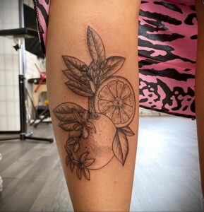 Фото тату апельсина, разрезанного пополам, с цветами и листьями, выполненной в технике графики на ноге. Детализированный рисунок с использованием теней.