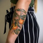 Фото реалистичной татуировки апельсиновой ветки с листьями и плодами на руке. Яркий рисунок татуировки с использованием оранжевых, зеленых и коричневых оттенков.