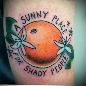 Фото татуировки на предплечье с изображением апельсина и надписью "A Sunny Place for Shady People". Выполнено в минималистичном стиле.