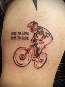 Фото тату на руке с изображением велосипедиста в красных тонах и надписью 'Ride to Live, Live to Ride'. Стиль татуировки - минимализм с элементами акварели.