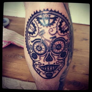 Фото тату на икре с изображением черепа в стиле сахарной черепушки, внутри которого находится велосипед. Детализированный рисунок татуировки в черных тонах.