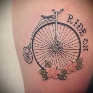 Фото тату: татуировка на бедре с изображением Penny-farthing (велосипед с большим передним колесом), украшенного цветами и надписью "Ride On".