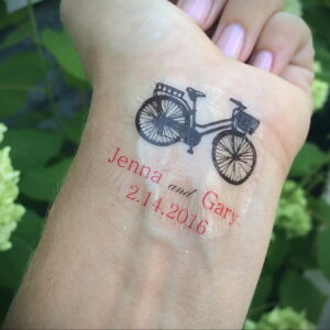 Фото женской татуировки с изображением велосипеда и надписью 'Jenna and Gary 2.14.2016' на запястье.