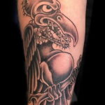 Фото тату грифа, держащего кости в лапах. Черно-белый рисунок в стиле blackwork с четкими контурами и минималистичной прорисовкой.