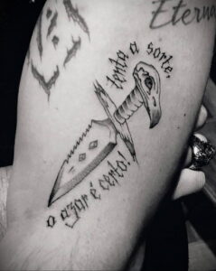Фото тату птицы, держащей в когтях кинжал, с латинской надписью, выполненное в черно-белой гамме с использованием тонких линий.