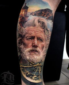 Фото тату на руке, изображающее портрет старого моряка с длинной бородой на фоне маяка и морского пейзажа. Реалистичный стиль и теплая цветовая гамма.