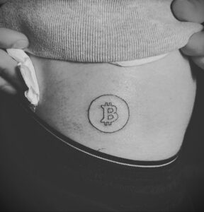 Черно-белое фото тату символа Bitcoin на пояснице. Минималистичный рисунок татуировки.