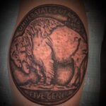Черно-белое фото татуировки на руке с изображением монеты Buffalo Nickel с изображением бизона. Детализированный рисунок татуировки.