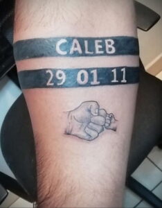 Фото тату на предплечье с надписью имени 'Caleb' и датой '29 01 11', заключенной в две черные полосы, под которой изображена стилизованная рукопожатие.