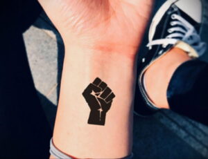Фото татуировки на запястье: силуэт сжатого кулака, выполненный в технике Blackwork, с добавлением небольшого символа внутри.