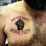Фото татуировки на груди с надписью "SAY HIS NAME GEORGE FLOYD" и изображением черного поднятого кулака. Татуировка выполнена в простом, но выразительном стиле.