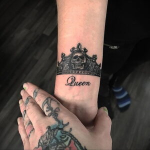 Фото тату: черная татуировка короны с черепом внутри и надписью 'Queen' на женском запястье. Детализированный рисунок татуировки в готическом стиле.
