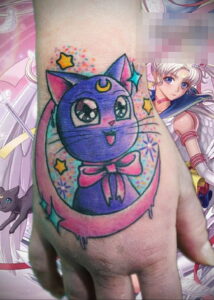 Татуировка на руке, изображающая кошку Луны из аниме Sailor Moon, окруженную звездами и ярким лунным фоном.