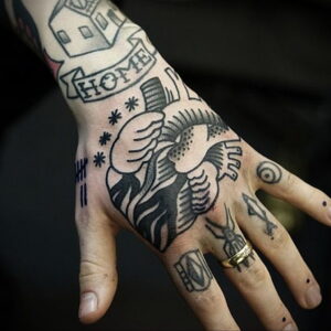 Фото тату: татуировка в стиле традишнл с изображением дома, сердца и надписи 'HOME', выполненная в черном цвете на руке.