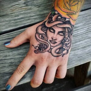 Черно-белое фото татуировки с изображением головы Медузы Горгоны на руке. Вокруг головы змеи, взгляд пронзительный.