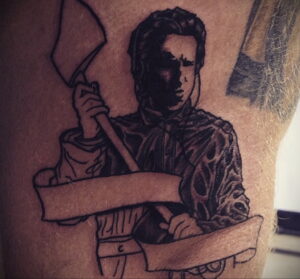 Фото татуировки с изображением Leatherface из фильма Техасская резня бензопилой, держащего бензопилу, выполненной в черно-белом стиле на плече.
