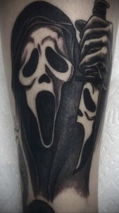 Черно-белая татуировка Ghostface с ножом на руке. Маска злодея изображена в динамичной позе, с ножом в руке.