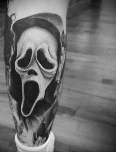Черно-белая реалистичная татуировка Ghostface на ноге. Маска злодея выполнена с высокой детализацией, видны все морщины и тени.