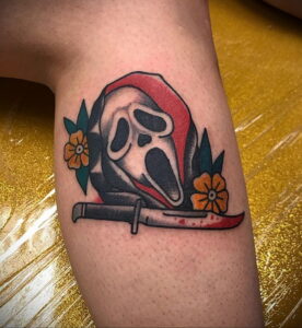 Татуировка Ghostface в стиле old school с добавлением цветов и ножа. Маска злодея окружена красными и синими цветами.