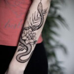 Черно-белое фото тату змеи, обвивающей букет цветов на женском предплечье. Детализированный рисунок с плавными линиями и реалистичными тенями.