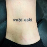 Фото минималистичной татуировки надписи "wabi sabi" на лодыжке. Простой и элегантный дизайн.