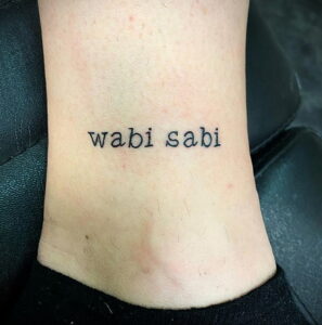 Фото минималистичной татуировки надписи "wabi sabi" на лодыжке. Простой и элегантный дизайн.