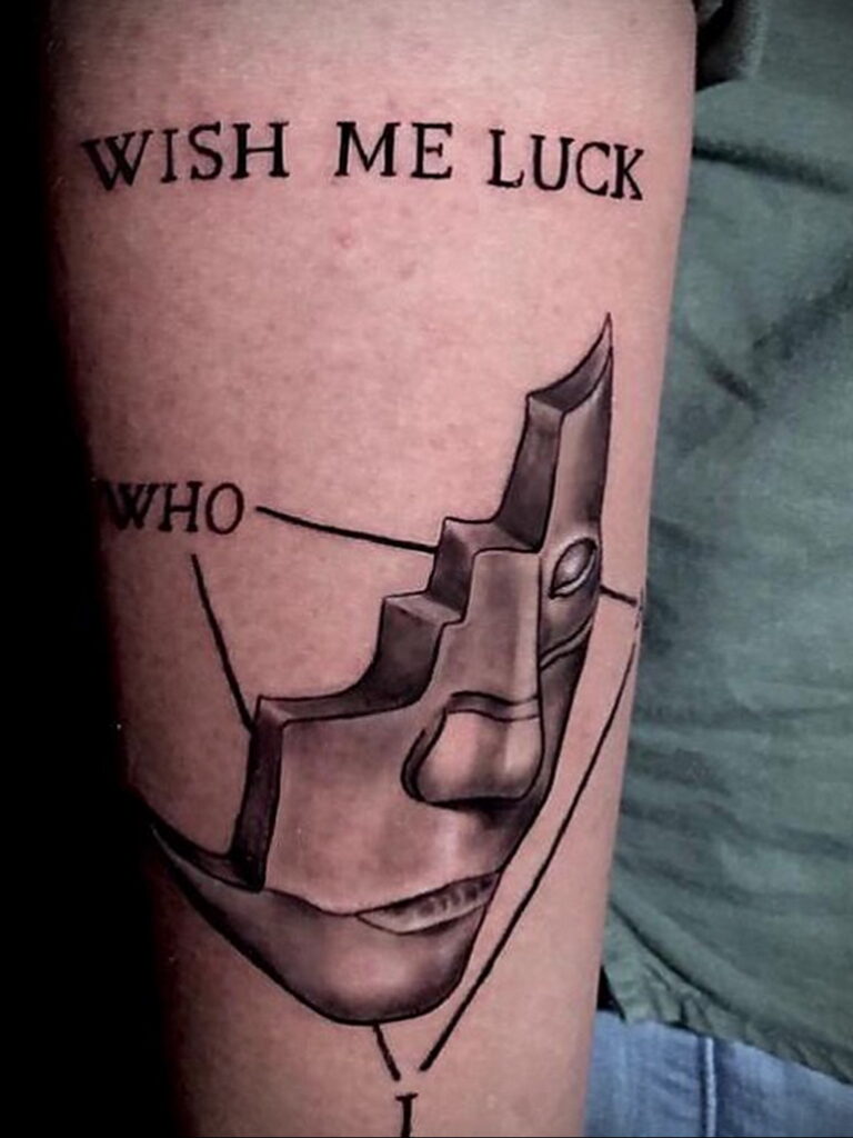 Фото тату на руке: стилизованное изображение маски и надпись 'Wish me luck'. Черно-белая татуировка в минималистичном стиле.