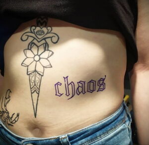 Фото тату на боку с изображением кинжала, цветка и надписи 'chaos'. Минималистичный рисунок татуировки в черном цвете.