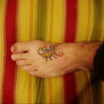 Фото небольшого цветного тату скорпиона на стопе. Простой и стилизованный рисунок, выполненный в ярких цветах. Подходит для тех, кто хочет незаметное и оригинальное украшение.