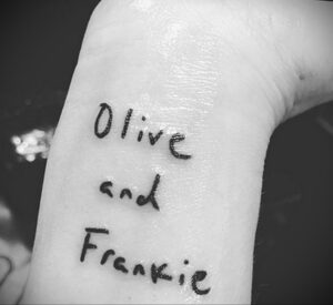 Черно-белое фото руки Париж Джексон с татуировкой, содержащей надпись "Olive and Frankie", выполненную от руки.
