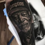 Фото тату: детализированный портрет рэпера Ice Cube в кепке Compton, выполненный в реалистичном стиле на предплечье, с эффектом дыма.