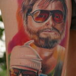 Фотография цветной татуировки с изображением персонажа The Dude из фильма "Большой Лебовски" в солнцезащитных очках и с ребенком в шапке. Яркий и детализированный рисунок.