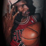 Фото тату с изображением Майкла Джордана в форме Chicago Bulls, показывающего два пальца. Детализированный портрет на предплечье.