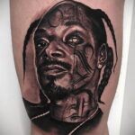 Черно-белое фото тату на руке, изображающее портрет Snoop Dogg. Детализированный рисунок с реалистичными тенями и текстурами кожи.