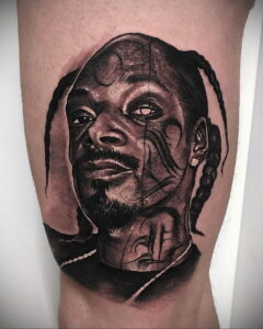 Черно-белое фото тату на руке, изображающее портрет Snoop Dogg. Детализированный рисунок с реалистичными тенями и текстурами кожи.