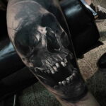 Фото тату: реалистичная черно-серая татуировка черепа с открытой пастью и острыми зубами на руке. Мрачный и детализированный рисунок.