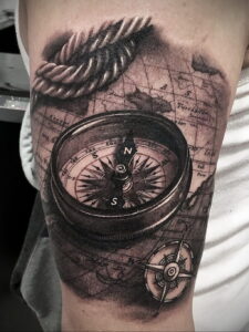 Фото тату с изображением компаса, карты и веревки на руке. Реалистичная проработка деталей и винтажный стиль.
