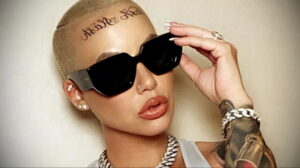 Amber Rose в черных очках с татуировкой 'Killa Kill' на лбу и татуировками на руке. Она держит очки и смотрит в камеру.
