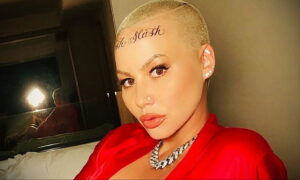 Фотография Amber Rose с выбритой головой и татуировкой 'Slash Slash' на лбу. Она смотрит в камеру, демонстрируя яркий макияж и украшения.