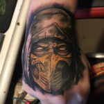 Фото татуировки Скорпиона из Mortal Kombat на руке. Желтая маска и детализированное лицо персонажа выделяются на фоне темного капюшона. Отличный пример для любителей видеоигр.