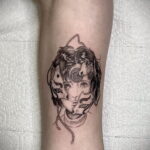 Фото тату на руке, изображающее лицо девушки, частично скрытое маской с гипнотическим узором. Детализированный черно-белый рисунок татуировки.