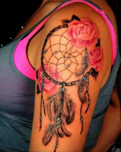 Фото тату: татуировка ловца снов с розовыми розами на плече. Черно-серая татуировка с акцентом на детализацию перьев и цветов.