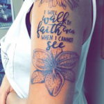 Фото тату: ромашка и надпись "I will walk by faith even when I cannot see" на руке девушки.