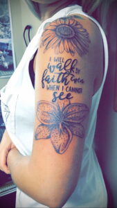 Фото тату: ромашка и надпись "I will walk by faith even when I cannot see" на руке девушки.
