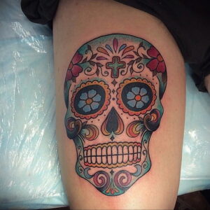 Фото тату: Цветная татуировка в стиле Sugar Skull с изображением украшенного черепа, расположенная на бедре.