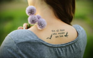 Фото тату: минималистичная татуировка с цитатой "Not all those who wander are lost" и изображением птицы на спине девушки.