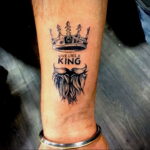 Фото тату: Татуировка с изображением короны, надписи 'Live Like a King' и бороды на руке мужчины. Черно-белый рисунок.