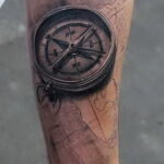 Фото тату: реалистичный рисунок компаса на фоне старинной карты, выполненный на предплечье. Черно-серые тона, детализированная проработка.